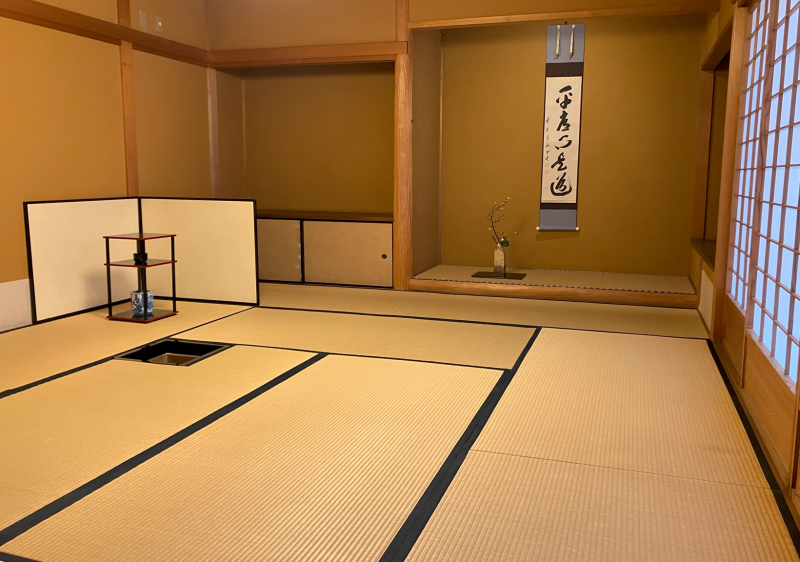 A large tea room, featuring tatami mats, shoji screens on the right, and in the back a tokonoma with a small flower arrangement and a hanging scroll hanging on the wall. To the left is a small standing byobu screen and a stand with tea utensils in front of a sunken hearth.
