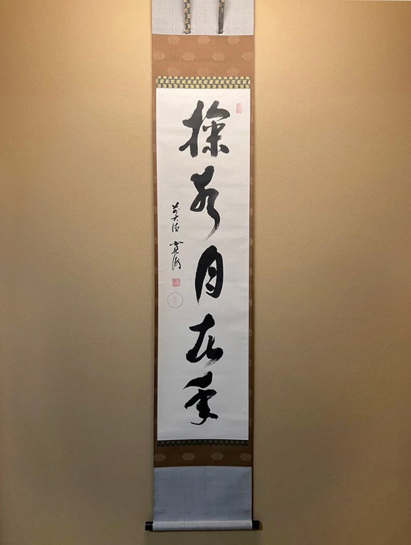 A vertical scroll with five Chinese characters brushed in black ink on white paper, mounted on brown and gold fabric, is hanging on a deep cream wall.