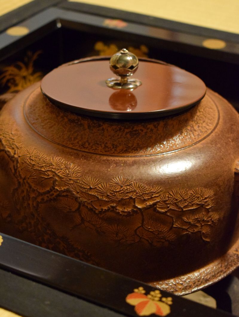 Large iron kettle, medium brown color with cast image of pine trees, sitting in a sunken hearth (ro), with its lid of shinier, smooth metal and silver knob ajar over the central mouth.