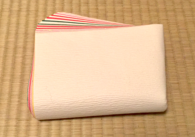 A folded stack of textured fabrics, white on the outside and with the edges of colored layers in pink and green showing from the artfully spread edges. The item is sitting on a tatami mat.