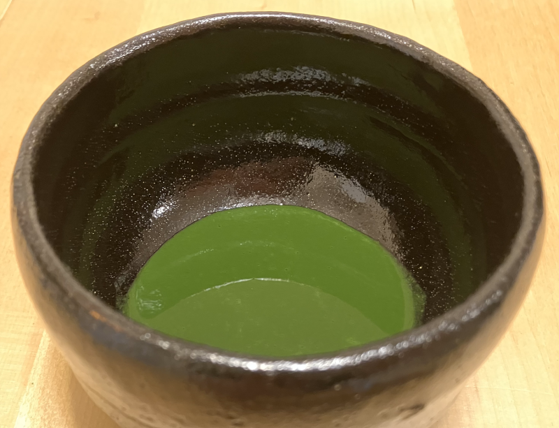 A blackish-brown ceramic bowl with thick tea inside that is dark green and smooth.