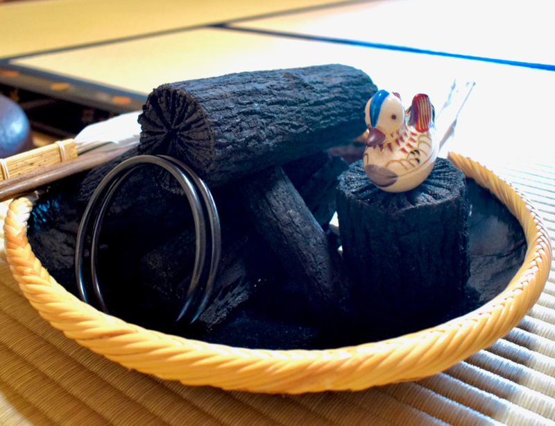 A neatly arranged pile of black charcoal in various shaoes and sizes sits in a shallow wicker basket. At the front are two round metal rings, for moving the kettle, and sitting on one piece of charcoal in the front right is a ceramic duck incense container. In the back a pair of metal chopsticks and a white father are just visible.