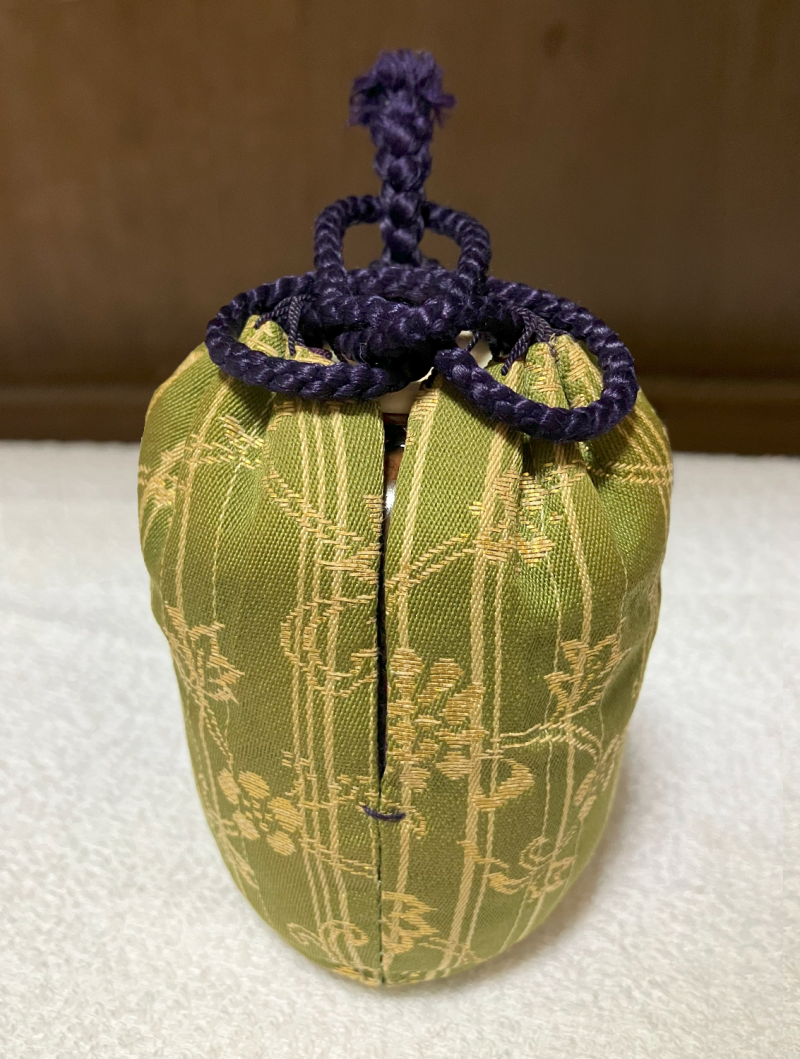 Green silk shifuku pouch with gold woven pattern of double line vines and flowers. The arrangement of the purple braided silk cord that closes the pouch in its empty indicates that the chaire inside is empty of tea.
