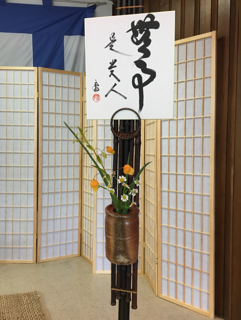 Calligraphed characters on rectangular paperboard hung from a pole with a small flower arrangement below. Behind are paper screens masking the room.