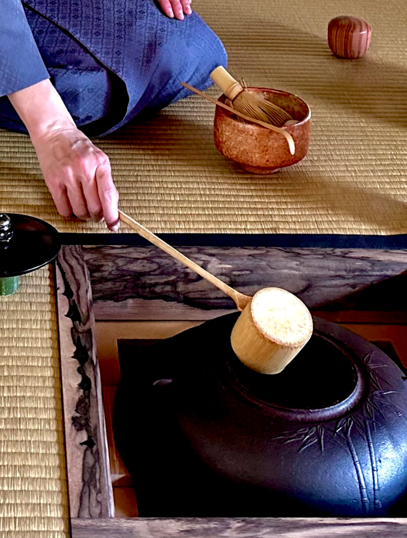 A man is seated on tatami with a teabowl in front of him, his hand placed on a ladle about to scoop water from the brazier in a sunken hearth.