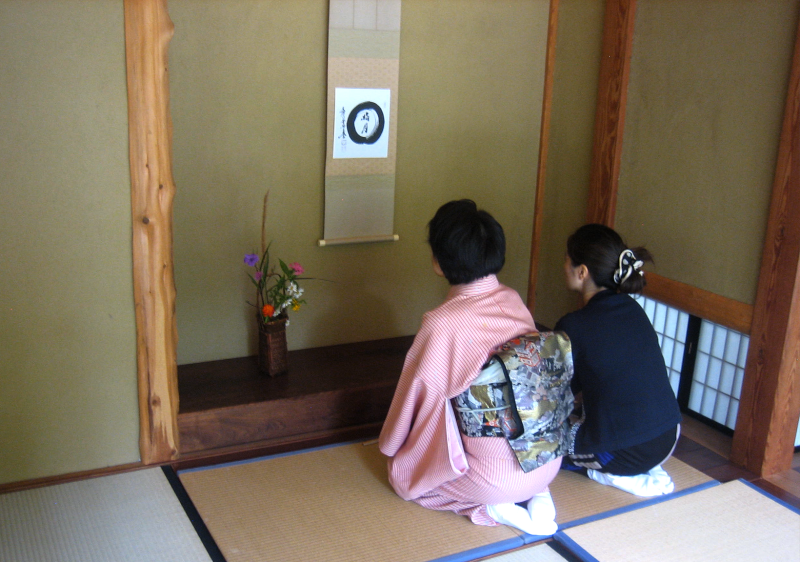 Two Japanese women, one in a pink kimono and elaborately-decorated obi and one in a dark blue shirt, kneeling on a tatami mat in front of an alcove that features a small jar with flowers and a hanging scroll with a circular pattern and calligraphy hung on the wall.