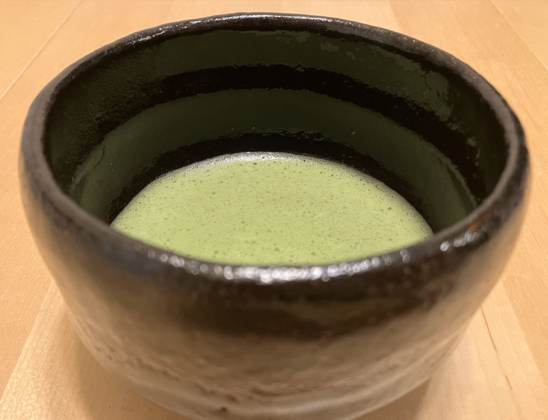 A blackish-brown ceramic bowl with thin tea inside that is light green and frothy.