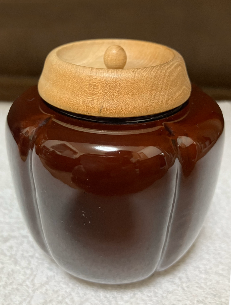 A shiny brown ceramic tea container with a carved wooden lid. It's shape is reminiscent of a bell pepper.