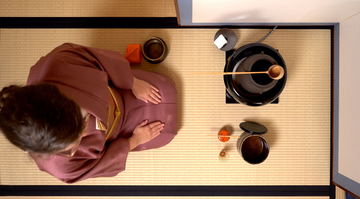 An overhead view of a woman in a muted dark pink kimono kneeling on tatami mats in front of a tea brazier and tea utensils.