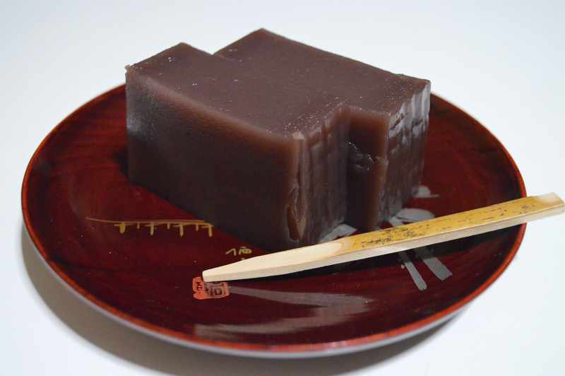 Closeup photograph of Mizu yōkan tea sweets, which are a rectangular cube shape with a deep purplish brown color. Two of them side by side sit on a red lacquer dish with a wooden stick next to them.
