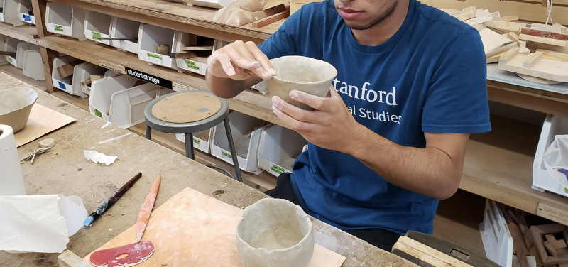 Photograph of a young man sitting at an art desk in a classroom, holding up a ceramic dish as he shapes the clay. Another clay bowl and sculpting tools rest on the table.