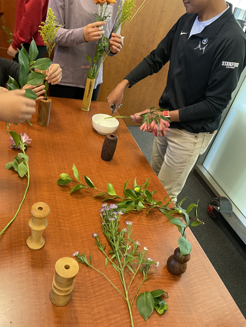 Photograph of a table laid out with ikebana stands and cuts of flowers and leaves for flower arrangement. Several students stand around preparing ikebana arrangements.