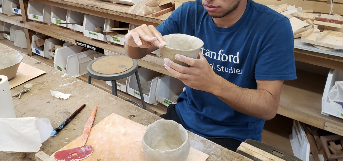 Photograph of a young man sitting at an art desk in a classroom, holding up a ceramic dish as he shapes the clay. Another clay bowl and sculpting tools rest on the table.