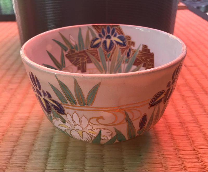 A white tea bowl with decorations of blue and white irises on the water with green leaves and wave patterns in gold, sitting on a tatami mat.
