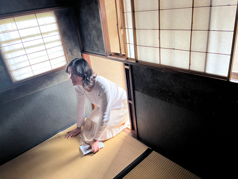 A young woman with medium-length black hair who is wearing white enters a tea room through its small square entrance, kneeling on the tatami.