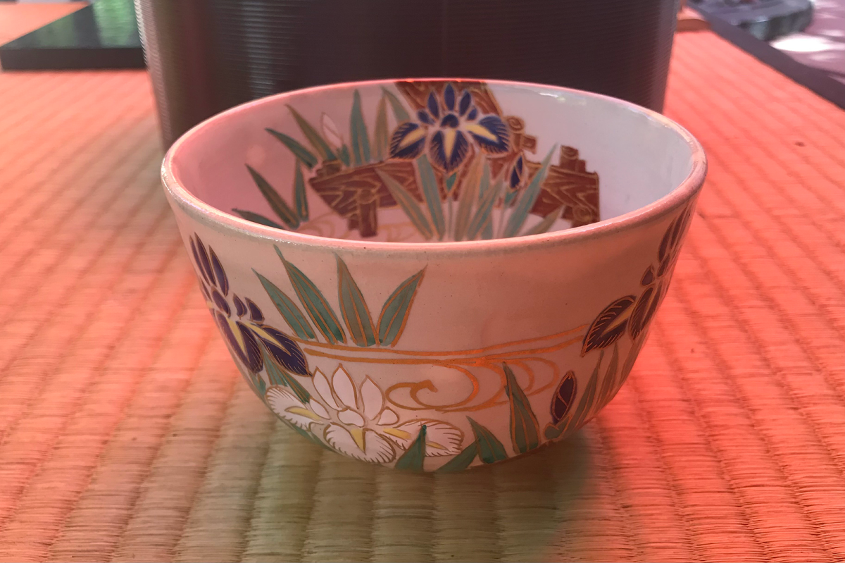A white tea bowl with decorations of blue and white irises on the water with green leaves and wave patterns in gold, sitting on a tatami mat.
