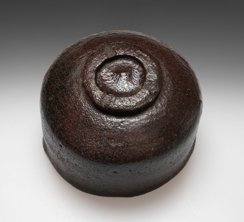 The bottom view of a black and red teabowl with a textured surface.
