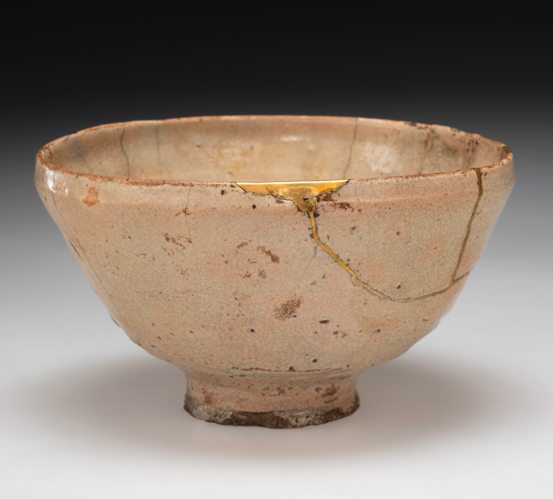 A light pinkish-tan ceramic tea bowl, showing a bit of age and repairs mended with gold kintsugi.